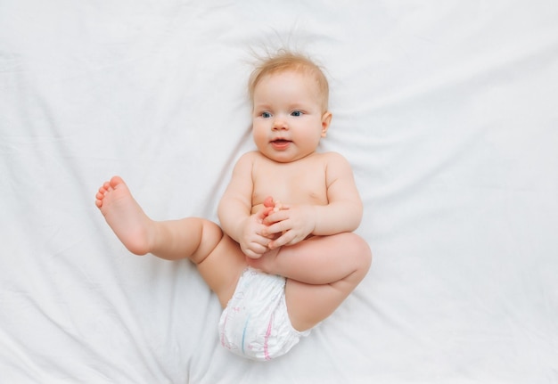 Ein fröhliches, fröhliches Baby in Windeln liegt auf einem weißen Bett und hält sein Bein Hochwertiger Fotoplatz für Text