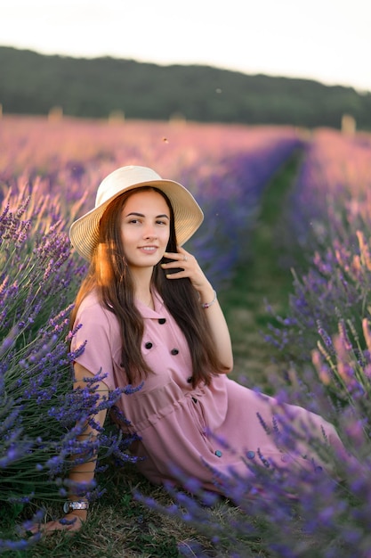 Ein fröhliches junges Mädchen in einem rosa Kleid und einem Strohhut in den Händen steht zwischen Lavendelbüschen