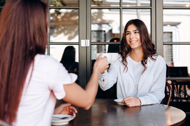 Ein fröhliches junges Mädchen und eine Freundin klirren an den Bechern. Sie sitzen im Café. Ansicht der ersten Person.