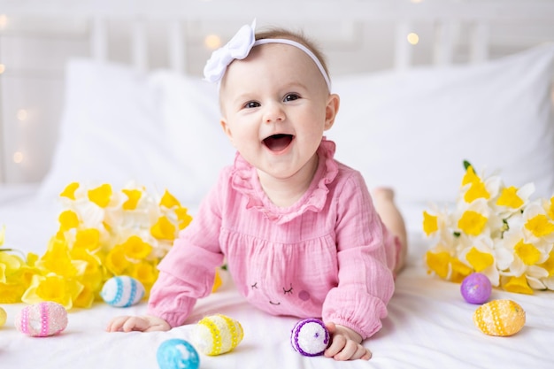 Ein fröhliches kaukasisches Mädchen mit einem Rand aus Hasenohren auf dem Kopf liegt zu Hause mit bunten Ostereiern und gelben Frühlingsblumen auf dem Bett und schaut auf die Kamera lacht Osterbaby