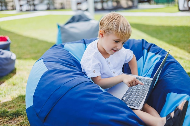 Ein fröhliches Kind spielt auf bunten Sitzsäcken auf der Straße Ein kleiner Junge sieht sich Zeichentrickfilme auf einem Laptop an, während er auf einem Stuhl im Park sitzt