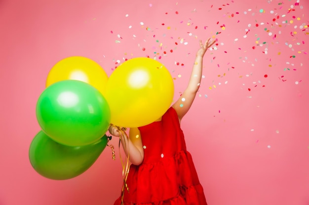 Ein fröhliches kleines Mädchen in einem roten Kleid feiert ihren Geburtstag und lässt buntes Konfetti auf einem rosa Hintergrund aus, ein Kind hält Luftballons und fängt Konfetti