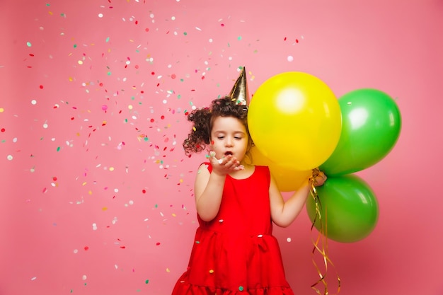 Ein fröhliches kleines Mädchen in einem roten Kleid feiert ihren Geburtstag und lässt buntes Konfetti auf einem rosa Hintergrund aus, ein Kind hält Luftballons und fängt Konfetti