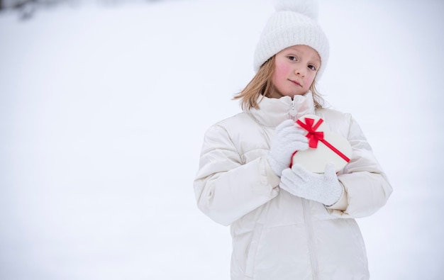 Foto ein fröhliches kleines mädchen in weißer winterkleidung steht vor dem schnee und hält eine herzförmige schachtel