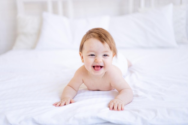 Ein fröhliches kleines Mädchen von sechs Monaten auf einem Bett in einer Windel krabbelt und lächelt ein kleines Kind auf einem Baumwollbett zu Hause das Konzept von Pflege und Hygiene