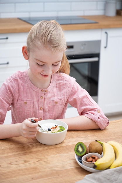 Ein fröhliches Mädchen in einem rosa Hemd sitzt am Küchentisch und isst Joghurt mit Müsli und frischem Obst Das Konzept der gesunden Ernährung