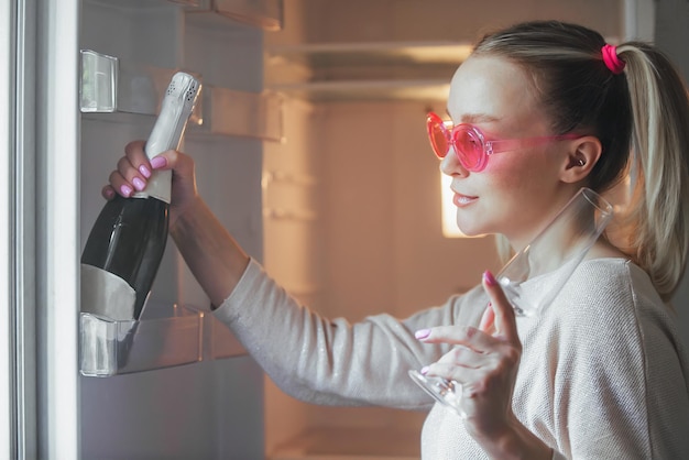 ein fröhliches mädchen mit rosa brille öffnete den kühlschrank und nimmt eine flasche champagner daraus