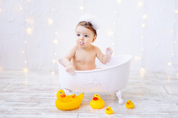 Ein fröhliches Mädchen von sechs Monaten badet in einem Bad mit Schaum und Gummienten, ein kleines Kind hat Spaß daran, mit Wasser zu spielen, das Konzept der Pflege und Hygiene