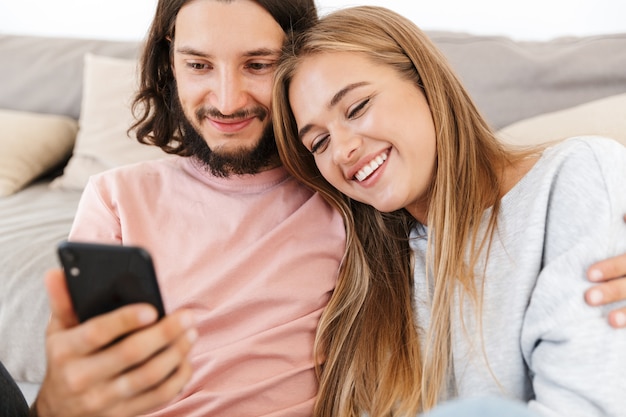 Ein fröhliches positives junges Liebespaar auf dem Sofa drinnen zu Hause mit dem Handy.