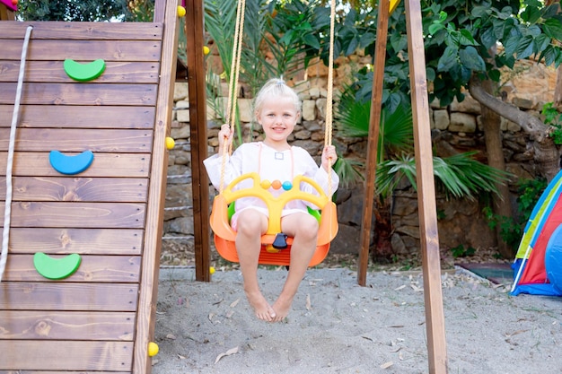 Ein fröhliches und fröhliches Kindermädchen schwingt im Sommer auf einer Schaukel auf einer Holzplattform und lächelt