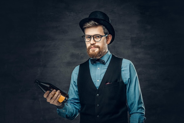 Ein funky bärtiger Hipster-Mann in einem blauen Hemd, einer eleganten Weste und einem Zylinder hält eine Craft-Bierflasche.