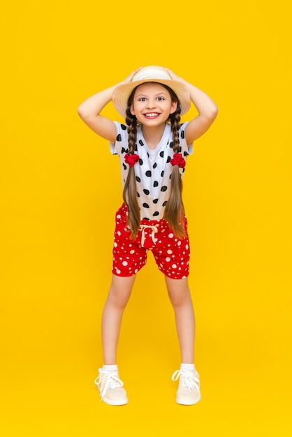 Ein Ganzkörperporträt eines kleinen Mädchens in roten Shorts, einem weißen T-Shirt und einem Strohhut, der ihren Hut mit beiden Händen hält und breit lächelt und sich im Sommer freut Gelb isolierter Hintergrund