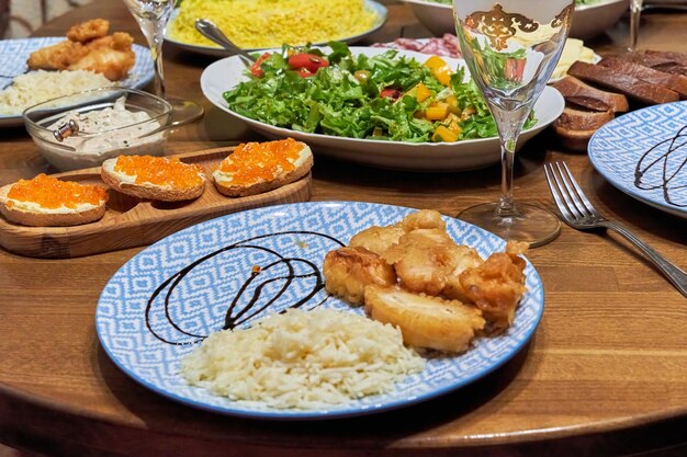 Ein gedeckter Esstisch mit verschiedenen Gerichten und Salaten, im Vordergrund ein Teller mit Reis und gebratenem Fisch