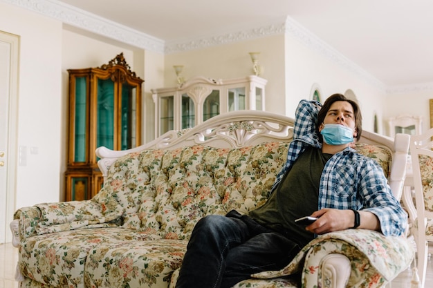 Ein gelangweilter, fröhlicher Mann in Quarantäne in einer medizinischen Maske sitzt auf einem Sofa und langweilt sich in Quarantäne des Coronavirus zu Hause. Pandemie COVID19