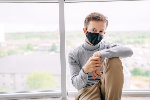 Ein gelangweilter Mann in Quarantäne, der eine medizinische Maske trägt, sitzt zu Hause und ist gelangweilt.