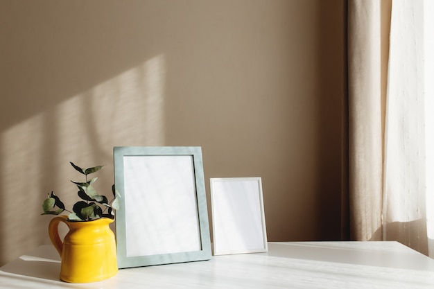 Ein gelber Keramikkrug oder eine gelbe Vase mit Eukalyptuszweigen, leere weiße Fotorahmen auf dem weißen Tisch im Inneren mit beigen Wänden nahe Fenster.