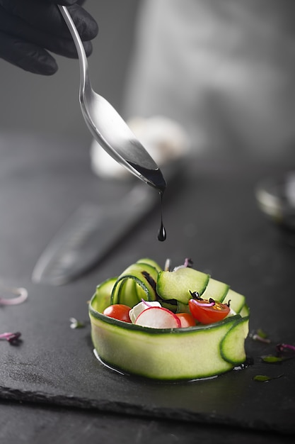 Ein Gemüsesnack aus frischem Gemüse wie Gurke, Tomate, Feta-Käse und Microgreens. Ein behandschuhter Koch gießt schwarze Soße über ein Gourmetgericht