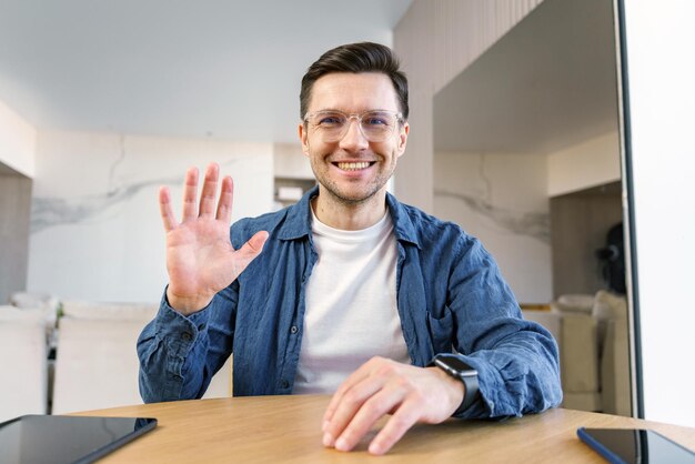 Ein genialer Mann winkt der Kamera mit einem Tablet und Laptop auf dem Tisch in einer modernen häuslichen Umgebung