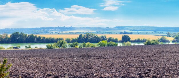 Ein gepflügtes Feld in der Nähe des Flusses an einem sonnigen Tag Ländliche Landschaft mit weißen Wolken am blauen Himmel