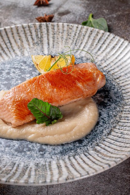 Ein Gericht aus der Speisekarte eines Restaurants servieren. Gebackenes Lachssteak mit Zitrone, Salat und Dill und Petersiliengrün auf einem Teller vor einem grauen Steintisch