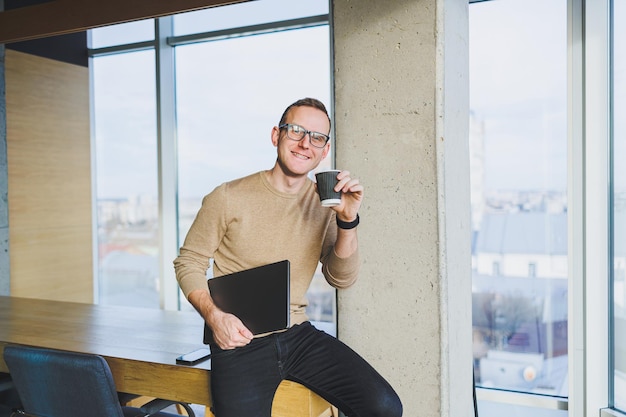 Ein Geschäftsmann mit Brille und stilvoller Kleidung trinkt Kaffee in einem modernen Arbeitsbereich Ein männlicher Freiberufler mit einem Laptop arbeitet aus der Ferne