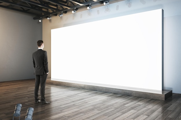 Foto ein geschäftsmann sucht nach einer beleuchteten leeren weißen plakattafel im innenraum mit holzfußboden und einem mock-up-platz für ihre anzeige