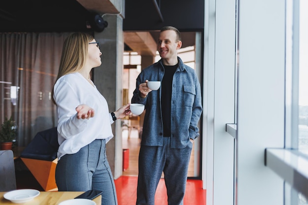 Ein Geschäftsmann und eine Geschäftsfrau trinken zusammen Kaffee und stehen vor den Fenstern eines Bürogebäudes mit Blick auf die Stadt. Zwei selbstbewusste Geschäftsleute, die in einem modernen Büro arbeiten