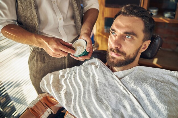 ein geschickter Barbier, der einen Rasierschaum für einen jungen Mann im Sessel mischt