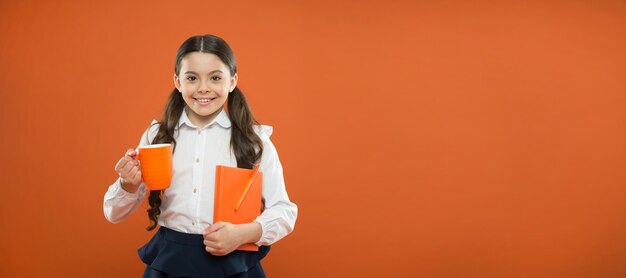 Ein gesunder Geschmack Nettes Mädchen mit Teetasse und Notizbuch auf orangefarbenem Hintergrund Kleines Schulkind, das Tee zum Frühstück trinkt Kleines Kind, das morgendlichen Tee genießt Kind mit Teepause in der Schule kopieren Raum