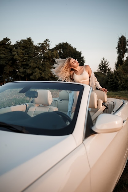 Ein glamouröses Luxusmädchen mit blonden Haaren, das beim Sitzen auf einem weißen Cabrio lächelt. Junge erfolgreiche Frau sitzt in ihrem weißen Auto