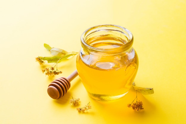 Ein Glas flüssigen Honigs von den Lindenblumen und ein Stock mit Honig auf gelber Oberfläche. Kopieren Sie Platz