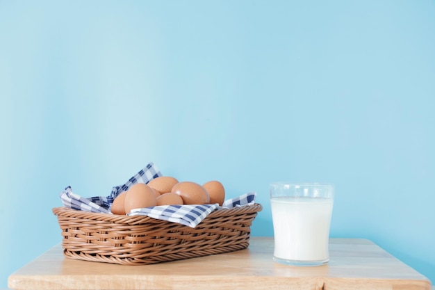 Ein Glas Milch und frische Eier im Korb auf einem Holztisch über einem blauen Hintergrund mit Kopienraum.