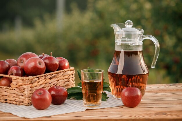 Ein Glas mit Apfelsaft und Korb mit Äpfeln auf Holztisch mit natürlichem Obstgartenhintergrund Vegetarische Fruchtzusammensetzung