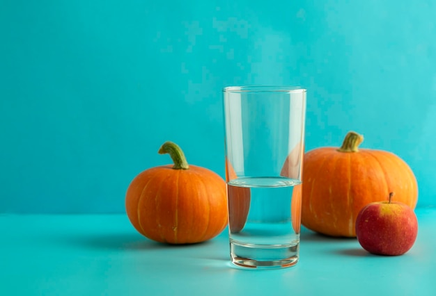 Ein Glas sauberes Wasser dahinter sind Kürbisse und ein Apfel auf blauem Hintergrund