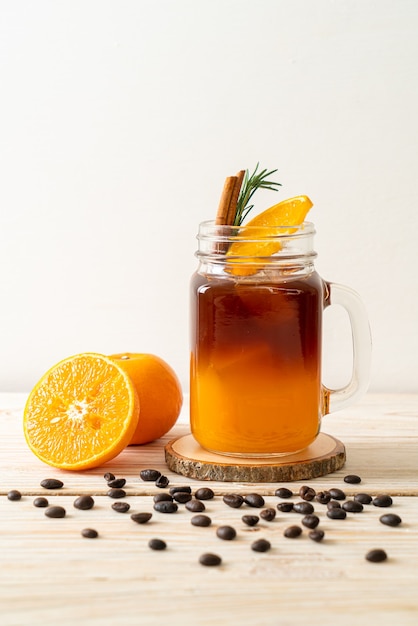 Ein Glas schwarzer Americano-Eiskaffee und eine Schicht Orangen- und Zitronensaft, dekoriert mit Rosmarin und Zimt auf Holzoberfläche