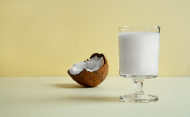 Ein Glas vegane Kokosmilch und ein Stück Kokosnuss auf hellgelbem Hintergrund