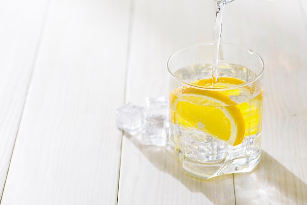 Ein Glas Wasser mit Zitrone und Eis auf einen weißen Tisch gießen