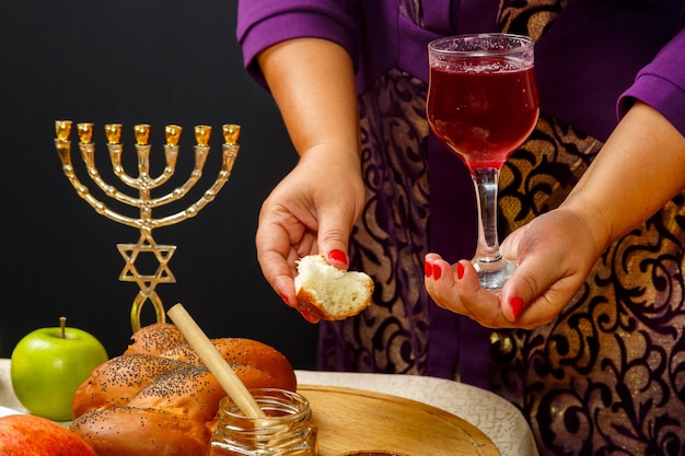 Ein Glas Wein und ein Stück Challah in Frauenhänden am Tag von Rosh Hashanah in Frauenhänden über einem Tisch mit Menorah