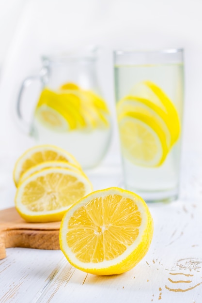 Ein Glasbecher und ein Krug kalte Limonade auf einem weißen hölzernen Hintergrund umgeben von Zitronen.