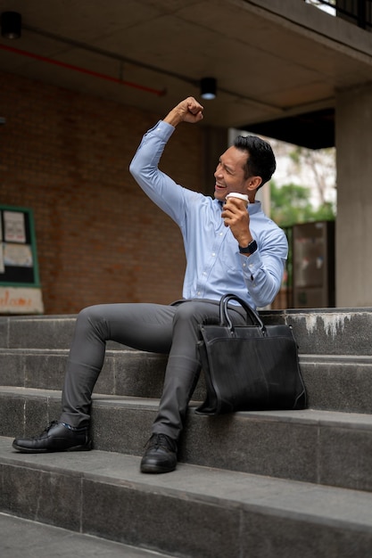Ein glücklicher asiatischer Geschäftsmann sitzt auf der Treppe mit einer Kaffeetasse und jubelt mit erhobener Faust