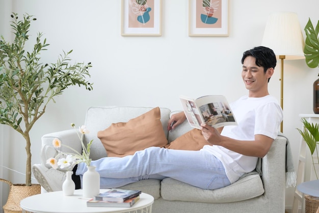 Ein glücklicher asiatischer Mann sitzt auf der Couch und liest ein Buch im Wohnzimmer zu Hause