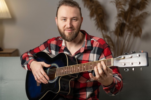 Ein glücklicher bärtiger Mann spielt Gitarre, während er abends zu Hause auf dem Sofa sitzt und eine sorglose Zeit zu Hause genießt