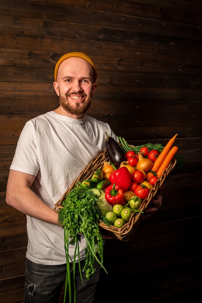 Ein glücklicher Bauer in einem weißen T-Shirt und Hut hält einen Korb mit frischem Obst und Gemüse