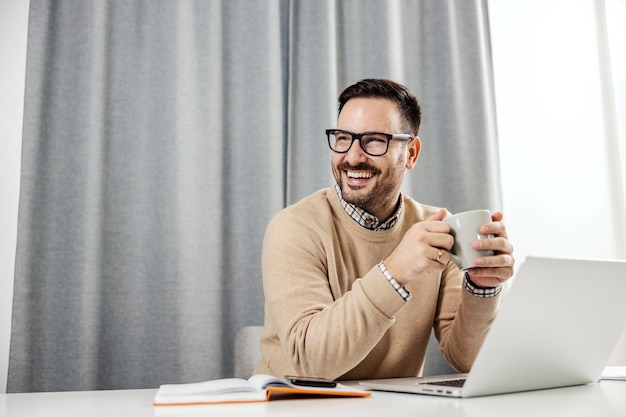 Ein glücklicher Geschäftsmann, der in seinem Heimbüro sitzt und eine Tasse Kaffee oder Tee hält und eine Pause macht