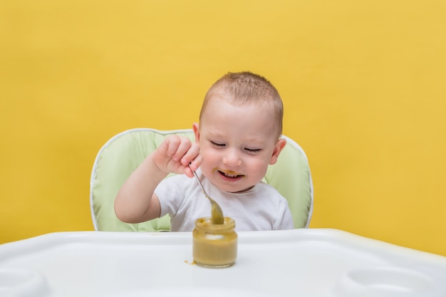 Ein glücklicher Junge isst allein Babynahrung an einem Tisch auf einem gelben isolierten