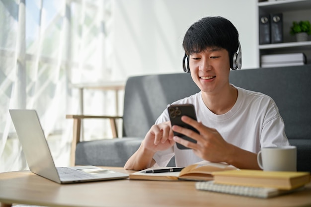 Ein glücklicher junger asiatischer Mann hört im Wohnzimmer Musik über seine Kopfhörer