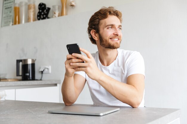 ein glücklicher junger bärtiger Mann am Tisch zu Hause, der per Telefon in der Nähe des Laptops spricht.