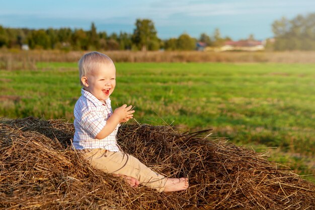 Ein glücklicher kleiner Junge, der auf einem Heuhaufen auf einem Feld sitzt und einen warmen Sommertag genießt