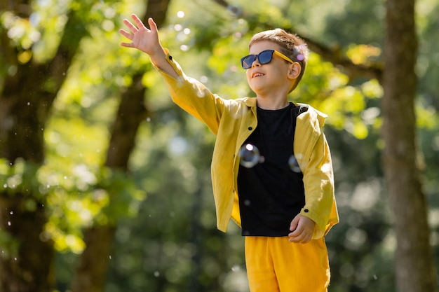 Ein glücklicher kleiner Junge mit Sonnenbrille fängt im Sommer Seifenblasen im Park