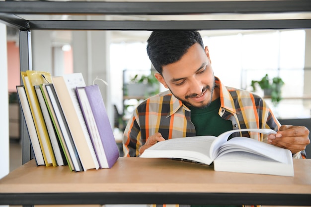 Ein glücklicher, kluger indischer oder arabischer Student mit gemischter Abstammung steht in der Bibliothek vor dem Hintergrund von Bücherregalen, hält viele Bücher in seinen Händen und blickt in die Kamera, lächelt freundlich